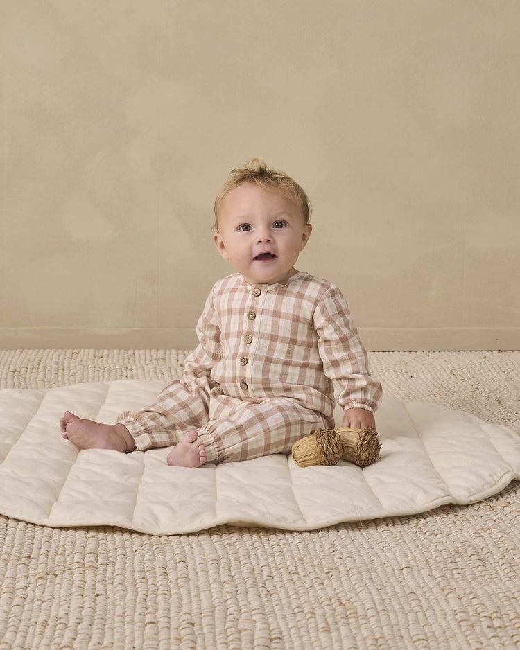 A baby wearing the quincy mae woven jumpsuit in rustic plaid sits on a cream quilted mat atop a textured rug, holding two small straw toys, with a neutral beige background.