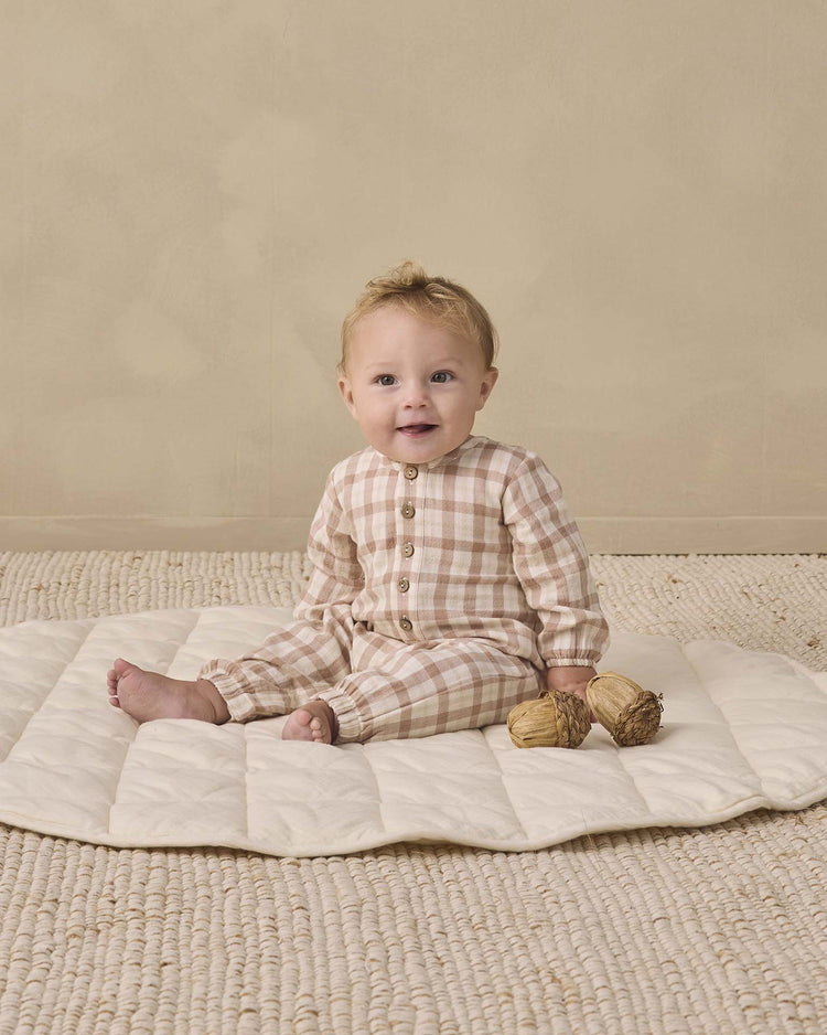 A baby wearing the quincy mae woven jumpsuit in rustic plaid sits on a quilted mat, holding two dried plants against a neutral background.