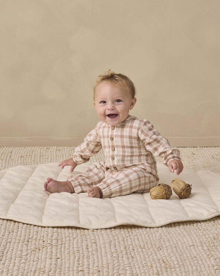 A baby wearing the quincy mae woven jumpsuit in rustic plaid smiles while sitting on a cream-colored mat atop a textured floor, with two decorative objects nearby against a beige background.