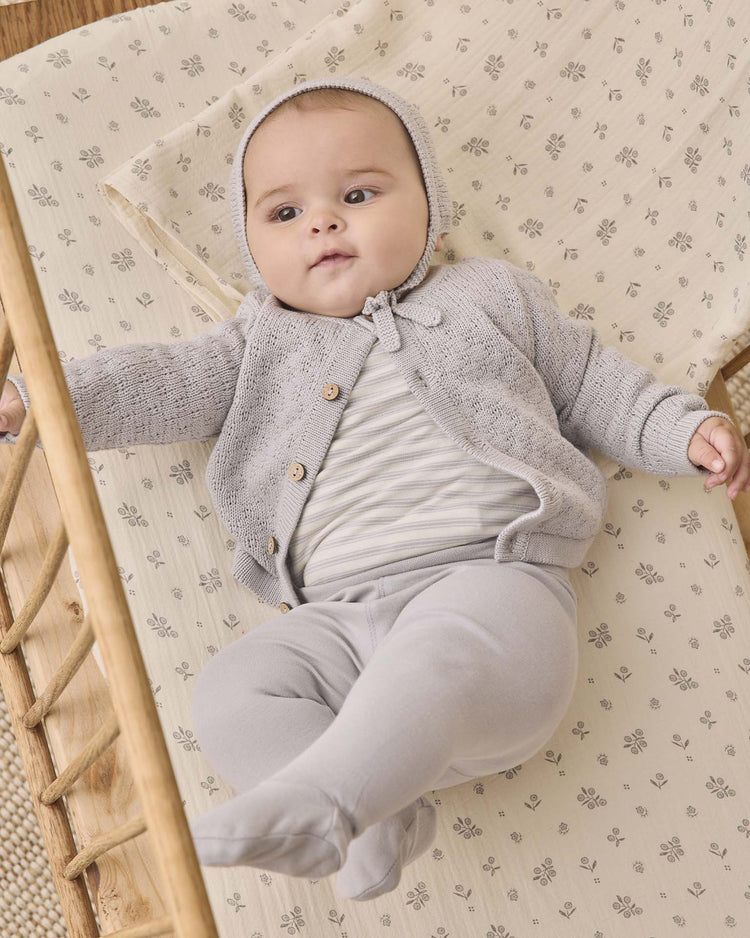 A baby wearing the Quincy Mae side snap bodysuit + pant set in cloud stripe lies on a patterned sheet in a wooden crib, looking up with one hand holding the crib side and legs outstretched.