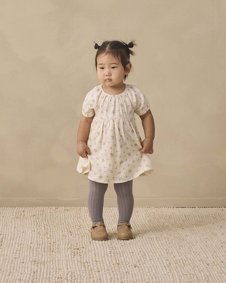 A young child with pigtails stands on a textured rug, wearing a light-colored dress with small prints, quincy mae tights in slate, and brown shoes, against a beige background.