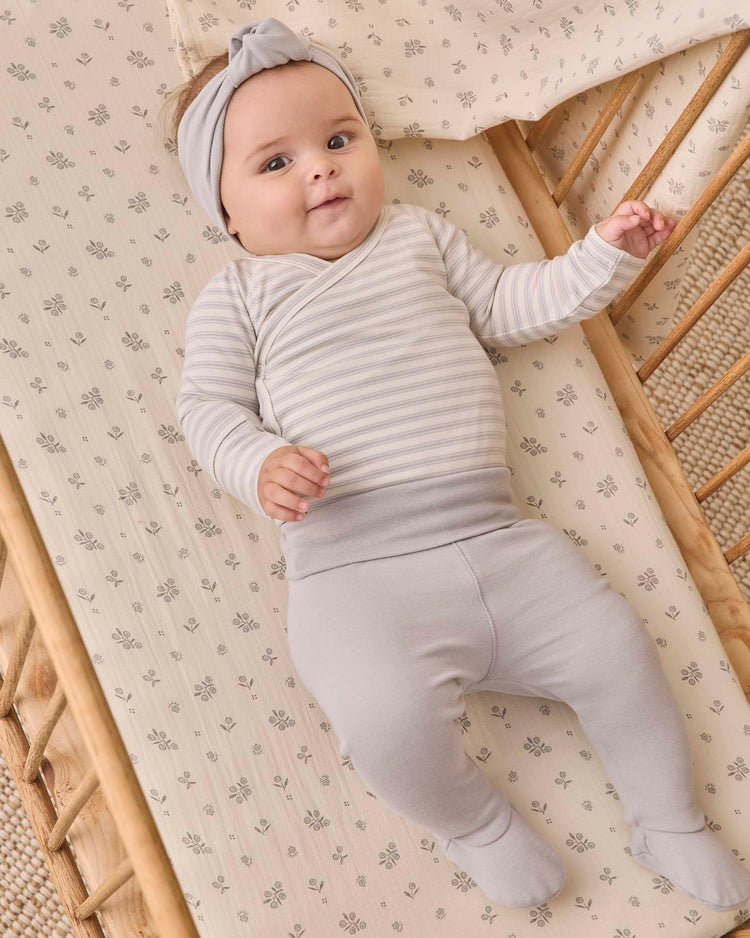 A baby wearing the quincy mae side snap bodysuit + pant set in cloud stripe, complete with a matching headband, lies in a wooden crib with floral bedding, looking up and smiling.