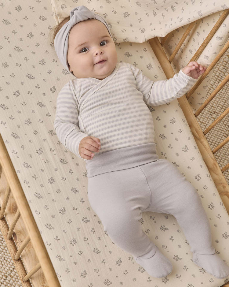 A baby wearing the quincy mae side snap bodysuit + pant set in cloud stripe, complete with a matching headband, lies in a wooden crib with floral bedding, looking up and smiling.