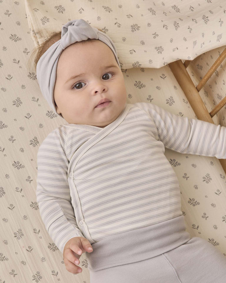 A baby wearing the quincy mae side snap bodysuit + pant set in cloud stripe, along with a gray headband, lies on a patterned crib sheet and gazes slightly upward with a calm expression.