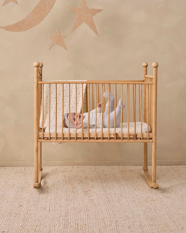 A baby dressed in the quincy mae side snap bodysuit + pant set in cloud stripe lies in a wooden crib on a textured rug, with star and moon decorations adorning the beige wall behind.