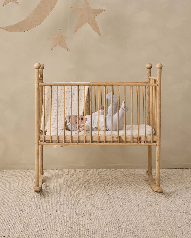 A baby dressed in the quincy mae side snap bodysuit + pant set in cloud stripe lies in a wooden crib on a textured rug, with star and moon decorations adorning the beige wall behind.