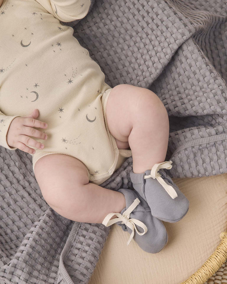 A baby wearing a cream onesie made from soft organic cotton, adorned with moon and star patterns, and cozy Quincy Mae baby booties in slate, lies peacefully on a textured gray blanket.