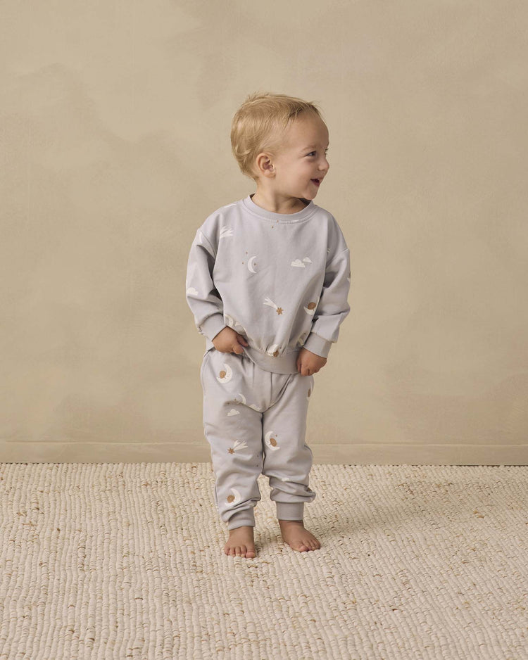 A light-haired toddler stands smiling on a textured rug, glancing to the side while wearing quincy mae’s relaxed sweatpant in sky friends—soft organic cotton sweatpants with a charming print of small clouds and moons, paired with a matching light gray sweatshirt.
