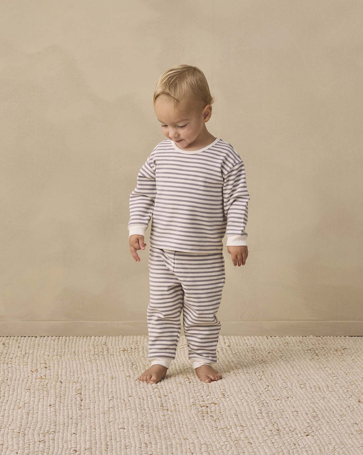 A toddler stands barefoot on a textured rug, dressed in the quincy mae cozy set in slate stripe—matching striped pajamas with drawstring pants. The child gazes downward with a slight smile against a plain beige background.