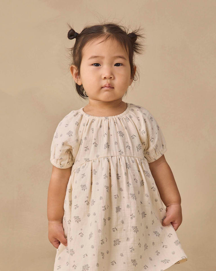 A young child with two small pigtails wears the quincy mae bella dress in winter berry, a cream-colored baby girl dress crafted from organic cotton and adorned with a delicate winter berry print. She stands against a plain beige background, looking slightly to the side.