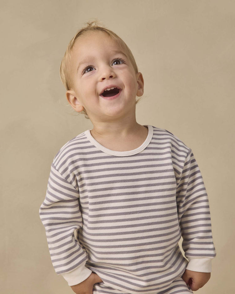 A smiling toddler in the quincy mae cozy set in slate stripe stands against a beige background, looking upward with hands tucked into the drawstring pants. Crafted from soft organic cotton, this outfit offers all-day comfort.