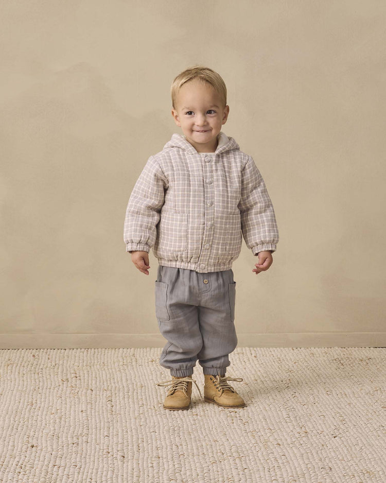 A small child stands smiling, wearing the quincy mae hooded woven jacket in blue gingham made from organic cotton, paired with gray pants with elastic cuffs and tan shoes, on a textured beige rug with a plain beige background.