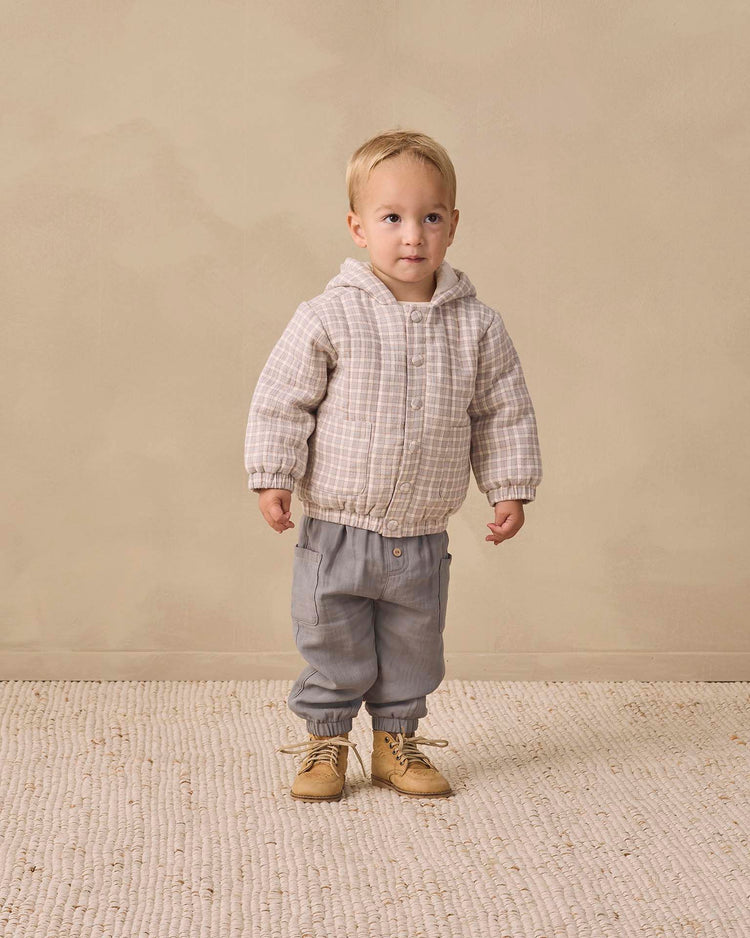 A young child stands indoors on a textured rug, wearing the quincy mae hooded woven jacket in blue gingham crafted from organic cotton, paired with gray pants and tan boots, against a beige background.