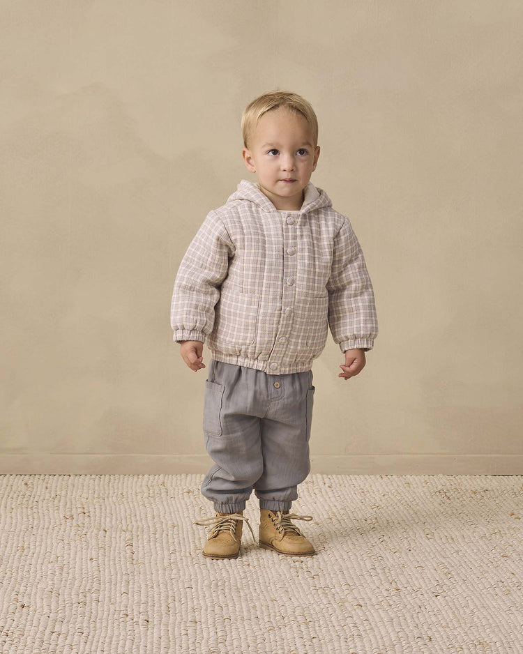 A young child stands indoors on a textured rug, wearing the quincy mae hooded woven jacket in blue gingham crafted from organic cotton, paired with gray pants and tan boots, against a beige background.