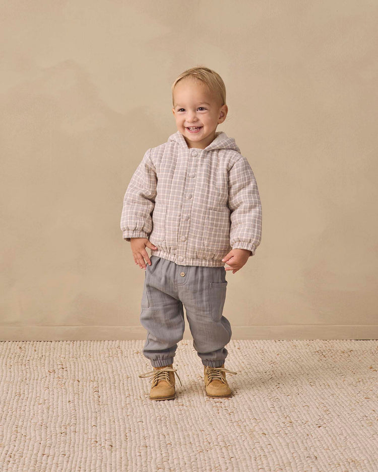A smiling toddler wears the quincy mae hooded woven jacket in blue gingham, paired with gray pants and tan boots, standing on a textured rug with a beige background.