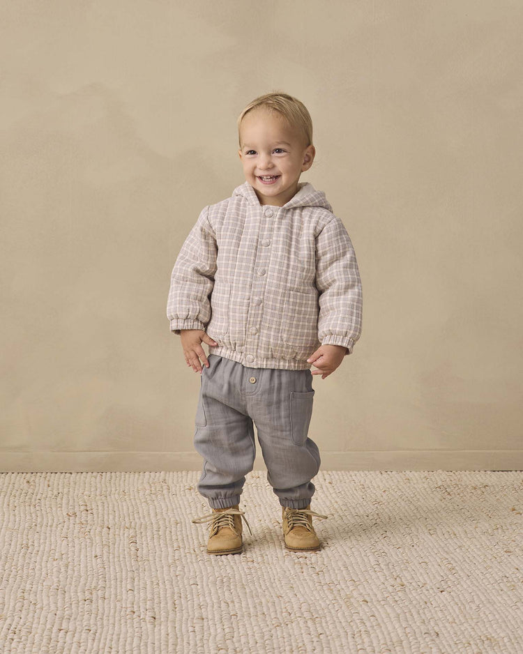A smiling toddler wears the quincy mae hooded woven jacket in blue gingham, paired with gray pants and tan boots, standing on a textured rug with a beige background.