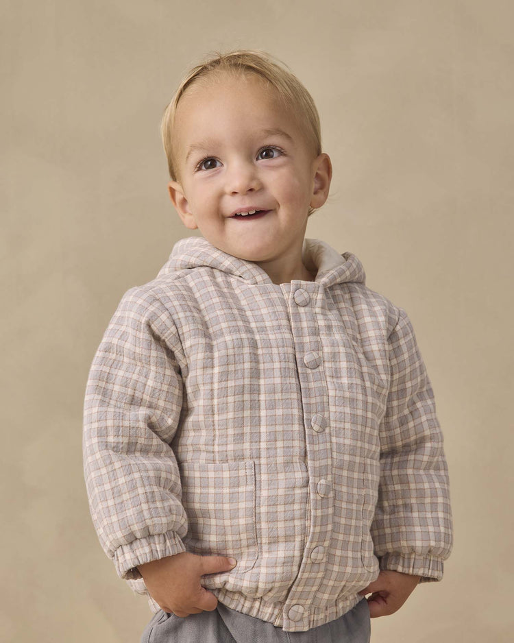 A young child with short blond hair smiles, wearing the quincy mae hooded woven jacket in blue gingham made from organic cotton, hands in pockets, standing against a plain beige background.