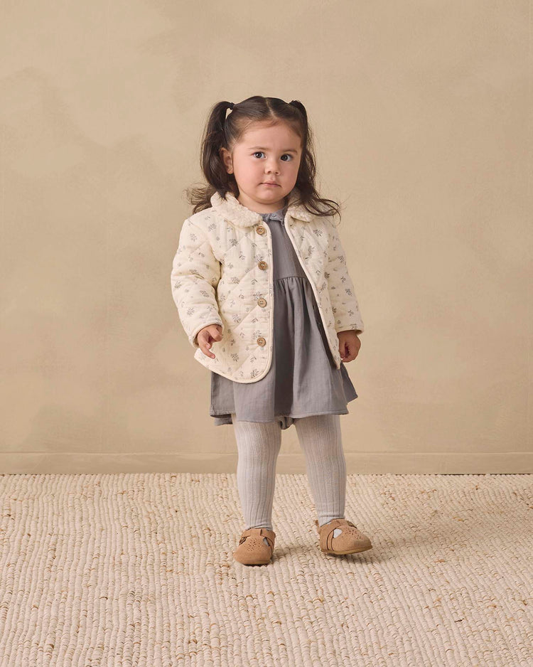 A young child with pigtails wears Quincy Mae's "tights in cloud," paired with a white patterned jacket, gray dress, and brown shoes, standing on a textured beige rug against a plain beige background.
