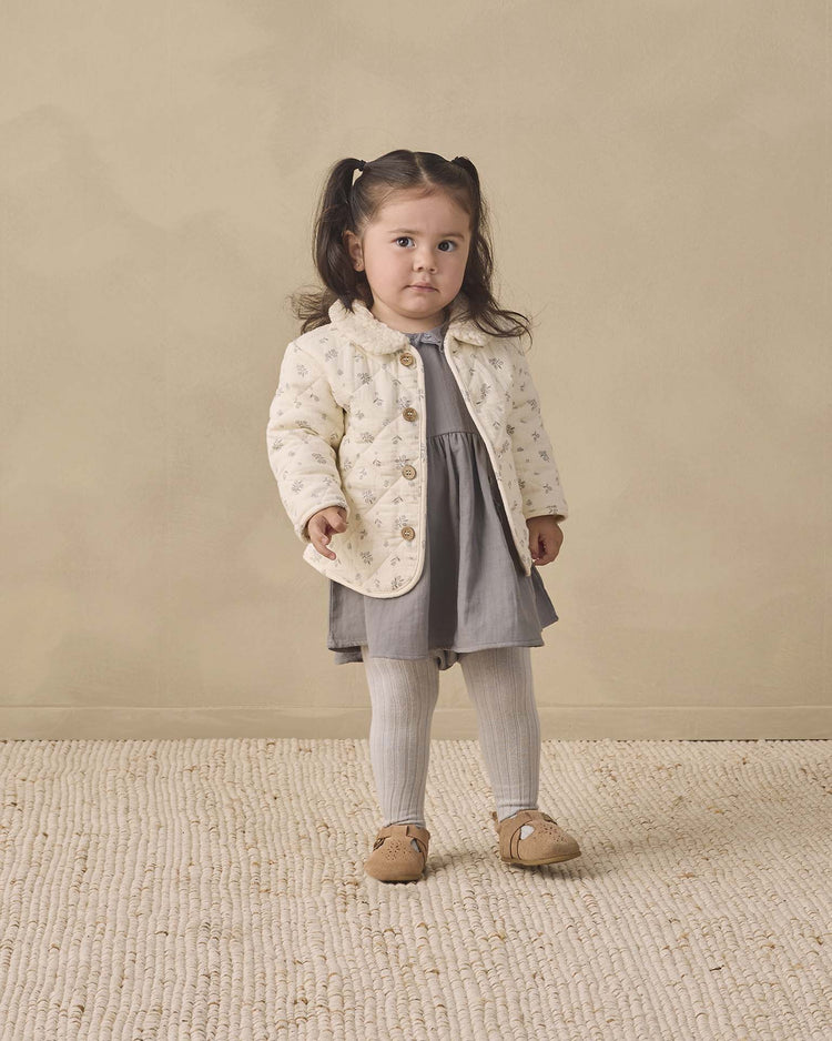 A young girl with dark hair in pigtails stands on a textured rug, wearing the quincy mae quilted jacket in winter berry—crafted from organic cotton and adorned with a delicate berry print—paired with a gray dress, light tights, and brown shoes, against a beige background.