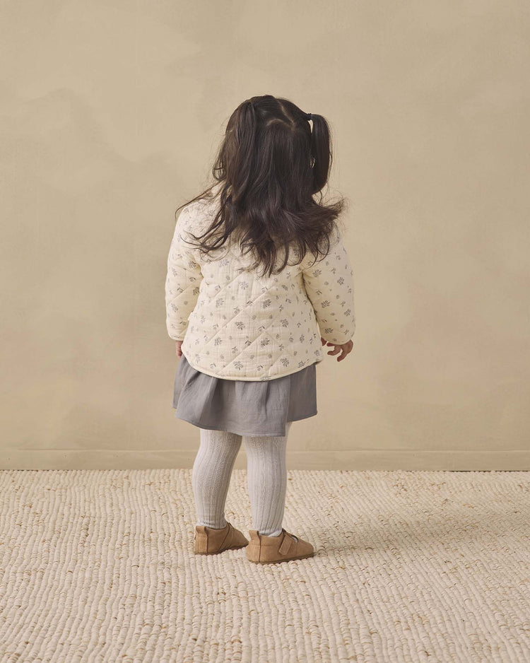 A young child with long dark hair in pigtails stands facing away, wearing the quincy mae quilted jacket in winter berry, along with a gray skirt, white tights, and brown shoes on a textured beige rug against a neutral background.