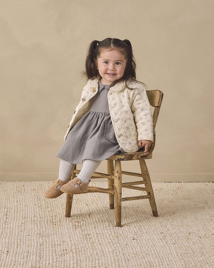A young girl with dark hair in pigtails sits smiling on a rustic wooden chair, wearing a gray dress, cream jacket, Quincy Mae tights in cloud made from organic cotton rib knit, and tan shoes, against a beige wall and textured rug.