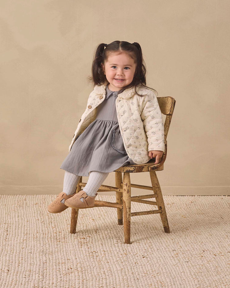 A young girl with dark hair in pigtails sits smiling on a rustic wooden chair, wearing a gray dress, cream jacket, Quincy Mae tights in cloud made from organic cotton rib knit, and tan shoes, against a beige wall and textured rug.