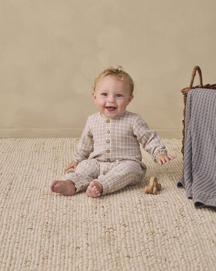 A smiling baby in the quincy mae woven jumpsuit in blue gingham sits on a textured rug next to a wooden toy and a wicker basket with a gray blanket draped over the side. A soft beige wall forms the background.