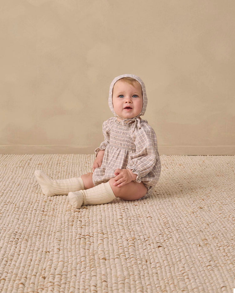 A baby in the quincy mae eden romper in blue gingham, paired with a bonnet and cream knee-high socks, sits on a textured beige rug against a plain light beige background, gazing slightly upward.