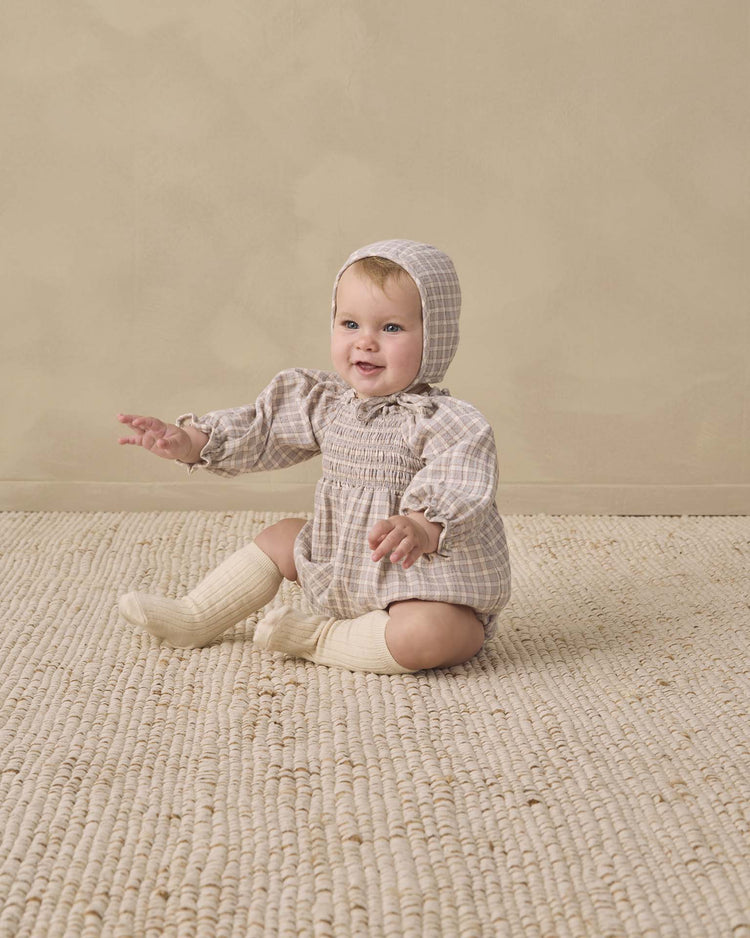 A baby wearing the quincy mae woven baby bonnet in blue gingham sits on a textured cream rug against a beige background, smiling with arms outstretched.