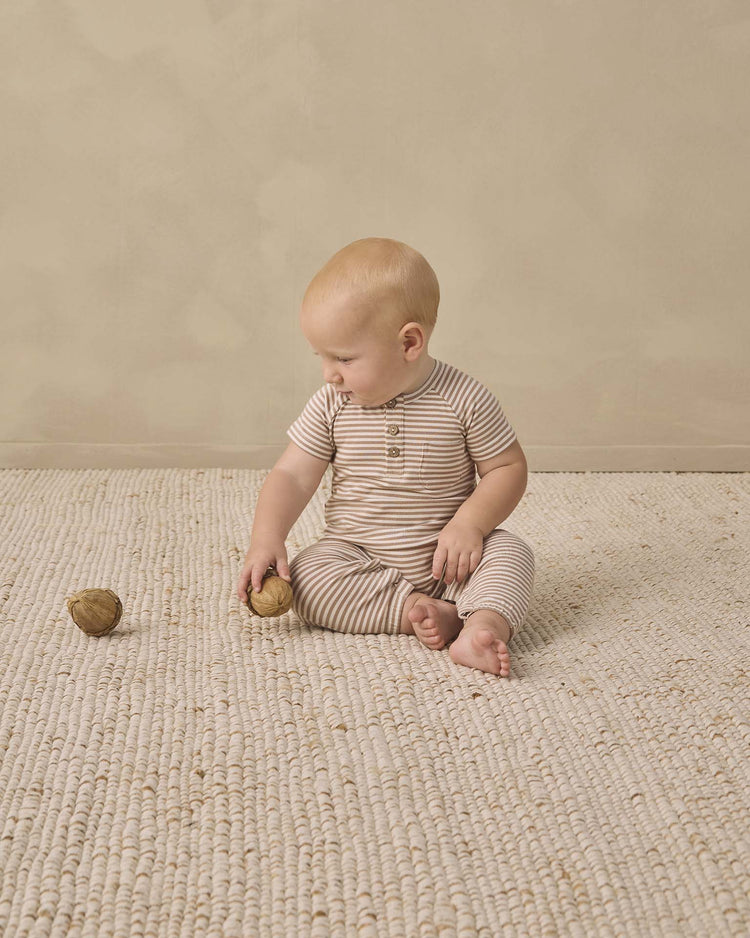 A baby wearing the quincy mae short sleeve jumpsuit in almond stripe sits on a textured cream rug, holding a walnut and gazing at another walnut on the floor. The background is plain beige.