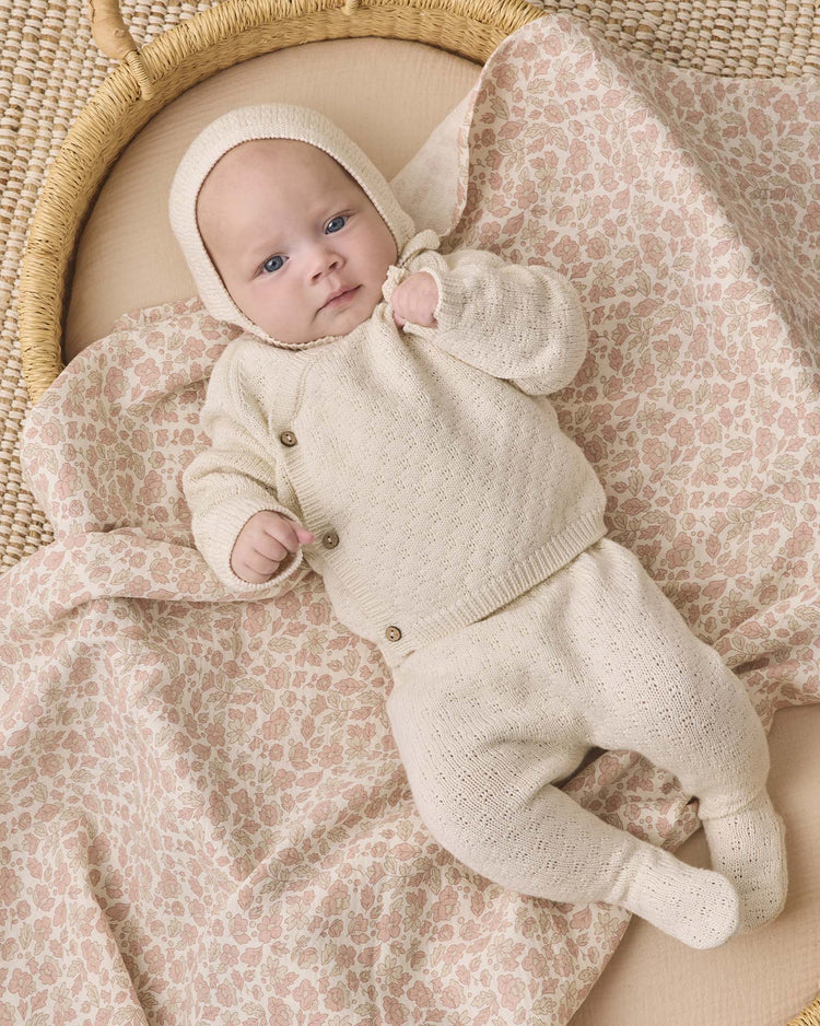 A baby dressed in the quincy mae knit wrap top + pant set in natural lies on a pink floral-patterned blanket inside a woven basket, gazing upward with one tiny fist raised. The organic cotton outfit brings an extra layer of comfort to this adorable moment.