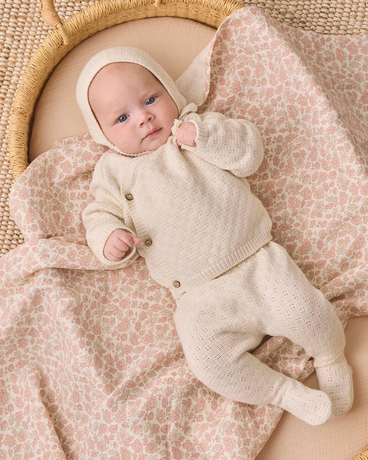 A baby dressed in the quincy mae knit wrap top + pant set in natural lies on a pink floral-patterned blanket inside a woven basket, gazing upward with one tiny fist raised. The organic cotton outfit brings an extra layer of comfort to this adorable moment.