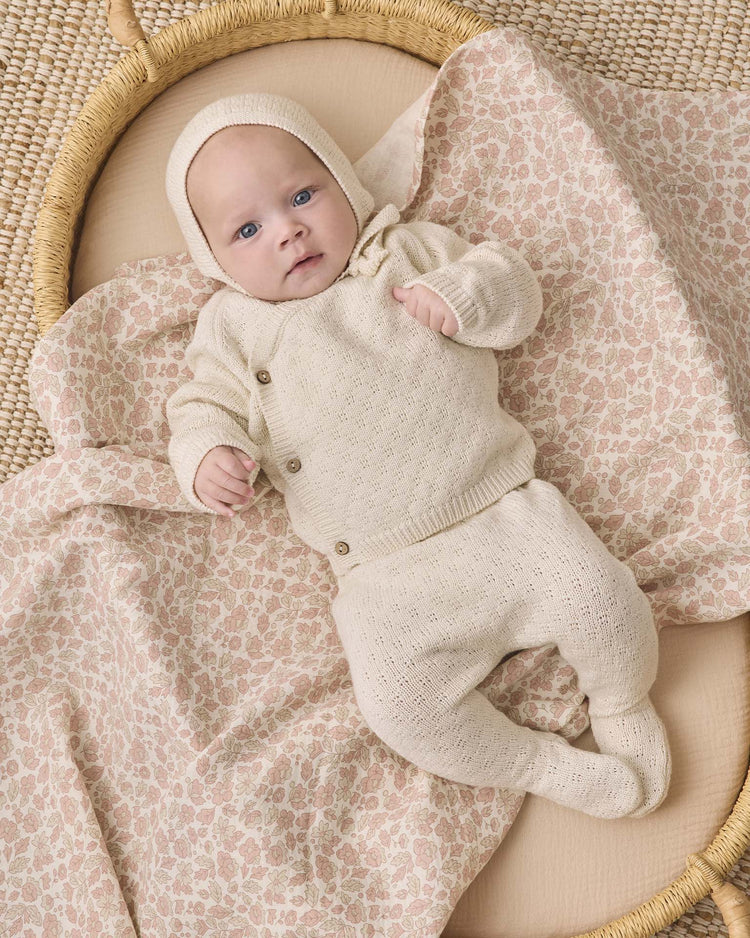 A baby dressed in the quincy mae knit wrap top + pant set in natural, complete with a cozy bonnet, lies on a soft, pink floral-patterned blanket in a woven basket, gazing up with blue eyes.
