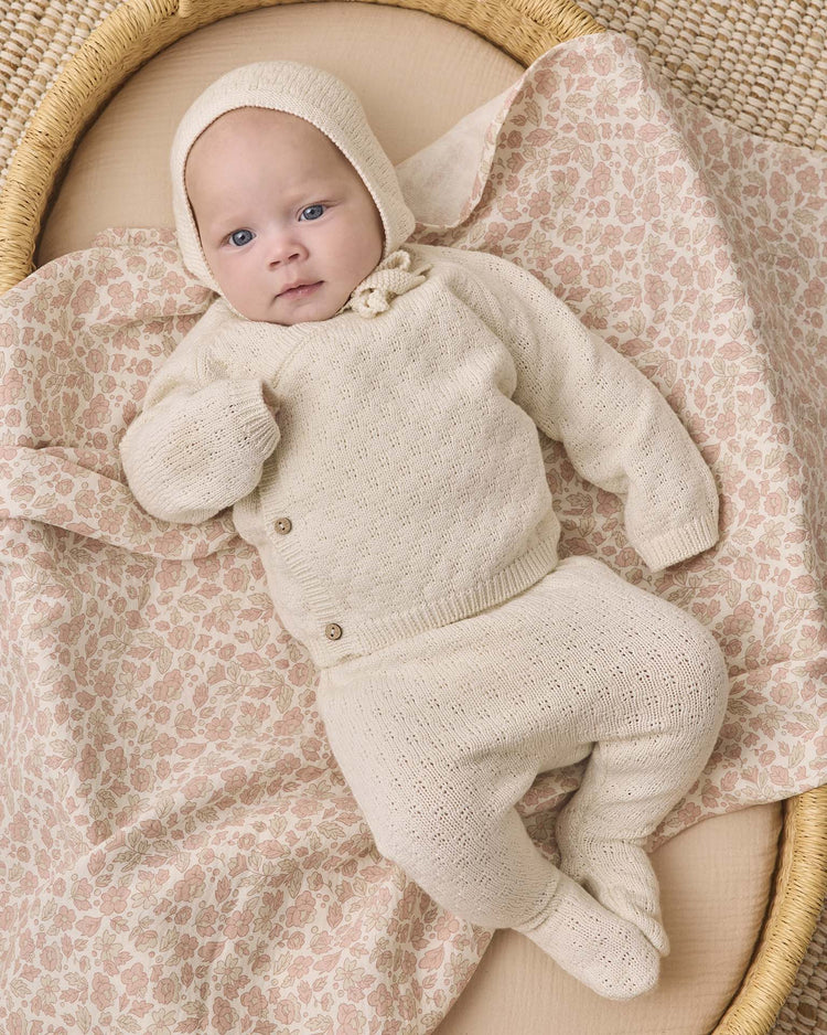 A baby in the quincy mae knit wrap top + pant set in natural, crafted from cozy cream organic cotton, lies on a floral-patterned blanket in a woven basket, gazing upward with a calm expression.