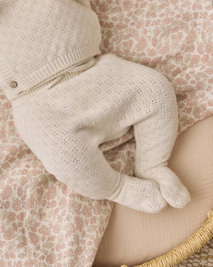 A baby wearing the Quincy Mae knit wrap top + pant set in natural lies on a floral-patterned blanket, with legs bent and feet touching each other.