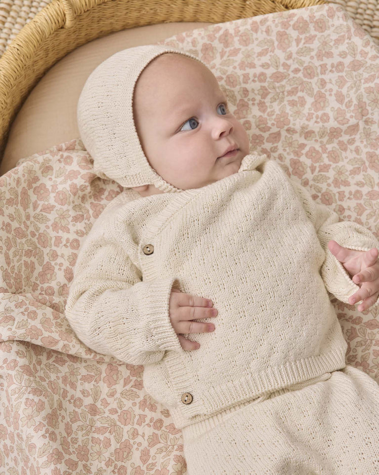 A baby wearing a quincy mae knit bonnet in natural and a cream knit outfit lies on a floral-patterned blanket in a wicker basket, looking to the side.