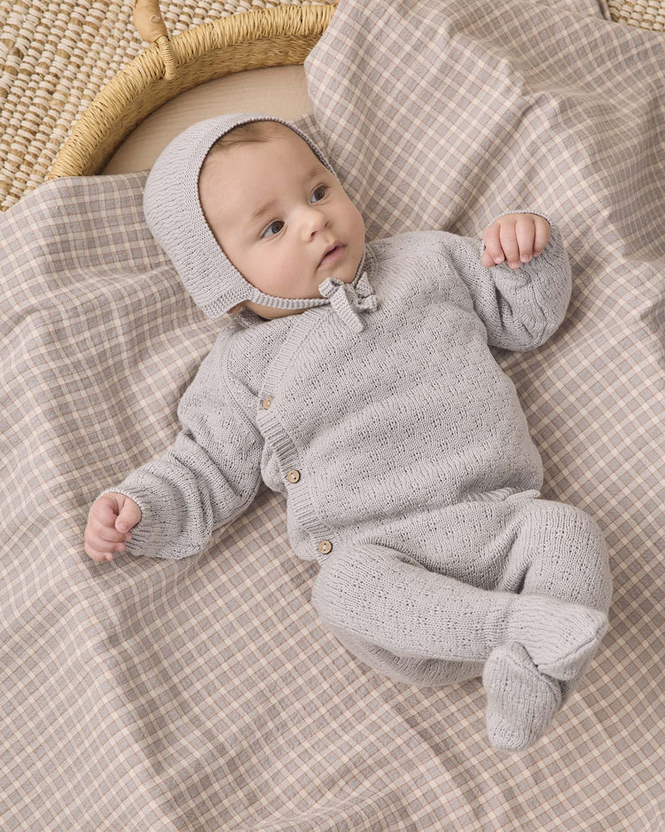A baby wearing the quincy mae knit wrap top + pant set in cloud and a matching bonnet lies on a beige and plaid-patterned blanket, looking off to the side with one arm raised.