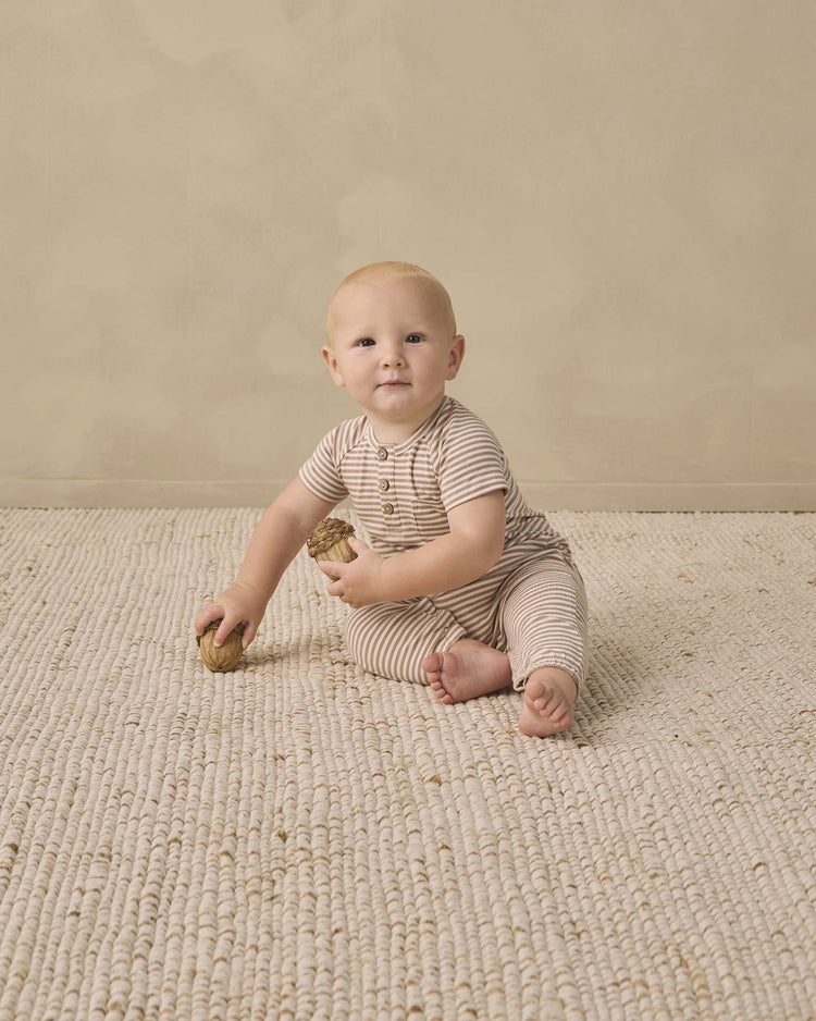 A baby wearing the quincy mae short sleeve jumpsuit in almond stripe sits on a textured beige rug, holding two wooden toys, with a neutral beige wall in the background.