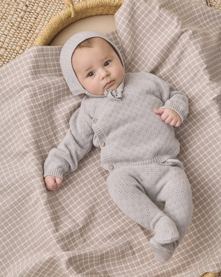 A baby wears the quincy mae knit wrap top + pant set in cloud, complete with a matching bonnet, while lying on a soft, beige plaid blanket beside a woven basket—a cozy scene perfect for showcasing organic cotton baby clothes.