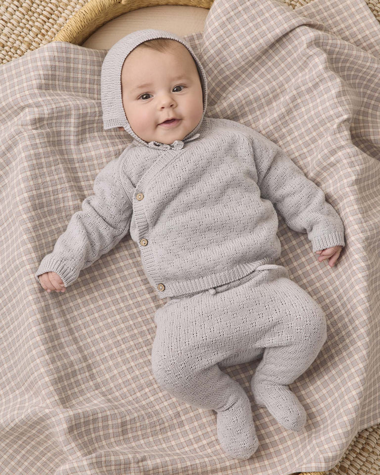 A baby wearing the quincy mae knit wrap top + pant set in cloud and bonnet lies on a beige and gray checkered blanket, looking up with a slight smile. This adorable newborn outfit is crafted from organic cotton for ultimate comfort.