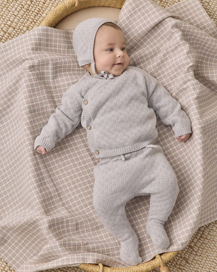 A baby wearing the Quincy Mae knit wrap top + pant set in cloud with a matching bonnet lies on a plaid blanket, looking off to the side and smiling gently.