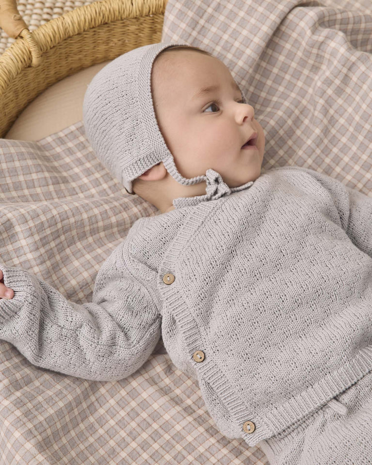 A baby wearing the quincy mae knit wrap top + pant set in cloud with a matching bonnet lies on a plaid blanket, looking to the side. A woven basket is partly visible in the background.
