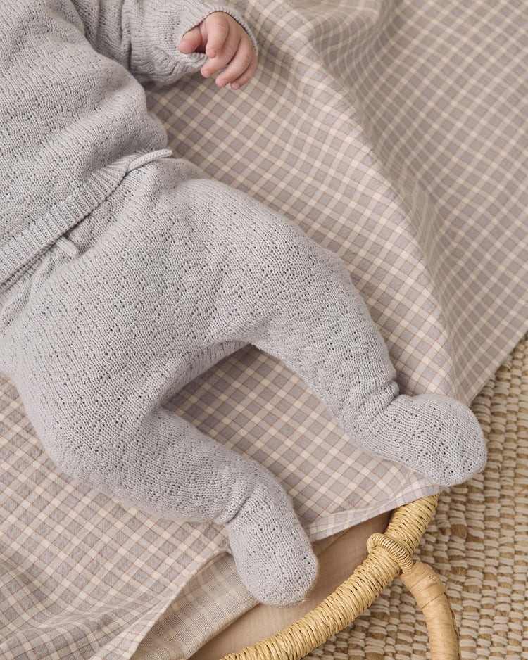 A baby wearing the quincy mae knit wrap top + pant set in cloud lies on a beige and pink plaid blanket in a woven basket, with only the lower body and left hand visible.