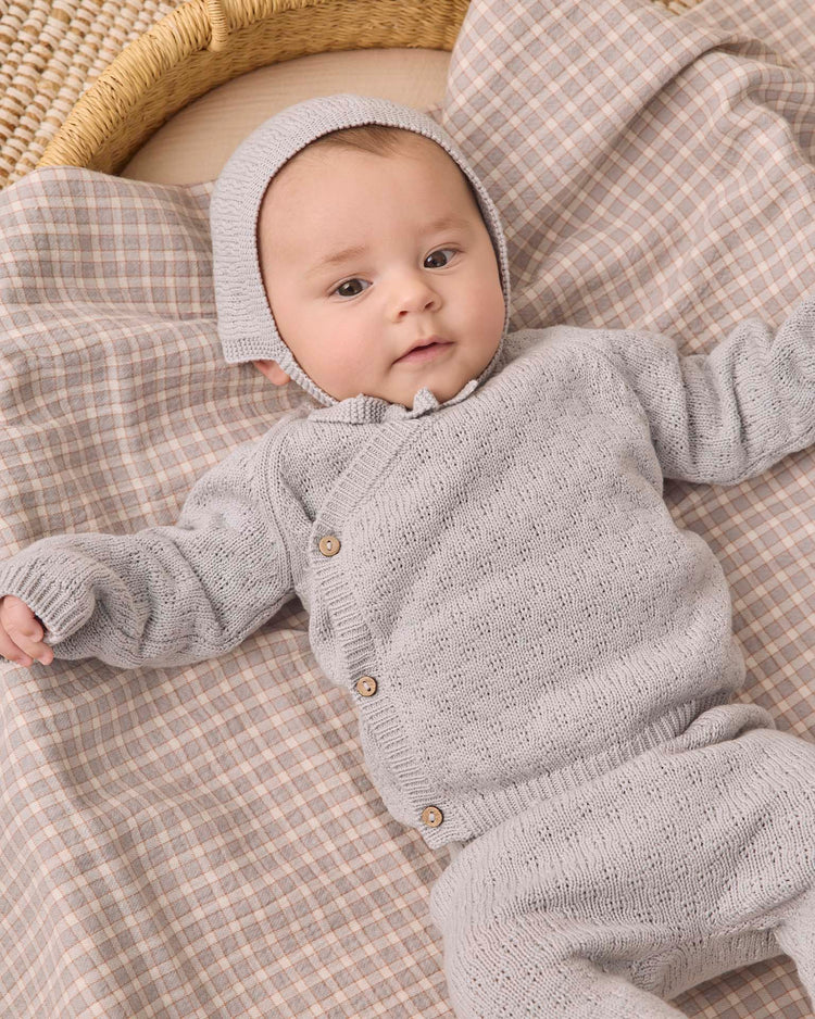 A baby wearing the quincy mae knit wrap top + pant set in cloud with a matching bonnet lies on a plaid blanket, looking up with arms outstretched.