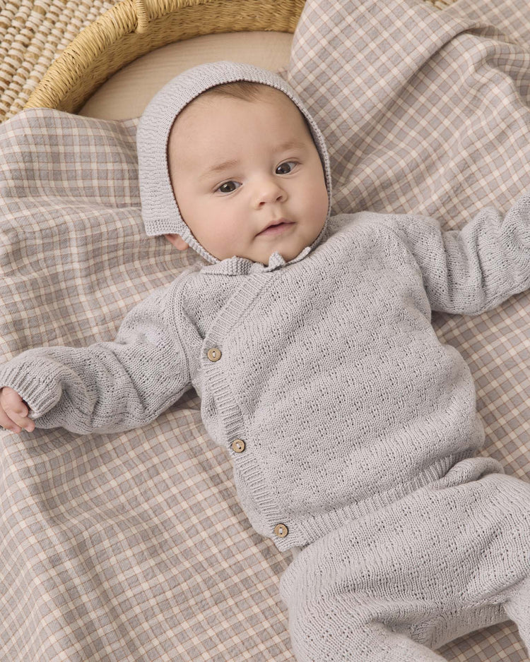 A baby wearing the quincy mae knit wrap top + pant set in cloud with a matching bonnet lies on a plaid blanket, looking up with arms outstretched.