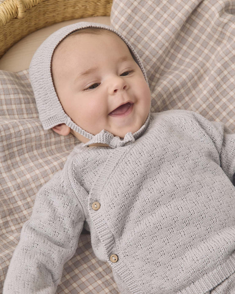 A baby wearing the Quincy Mae knit wrap top + pant set in cloud rests on a plaid blanket, smiling and looking up. The cozy ensemble is crafted from organic cotton, featuring soft knit footed pants that keep little toes warm and comfortable.