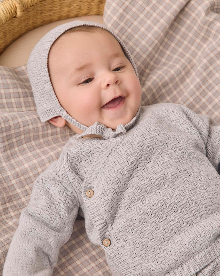 A baby wearing a gray knit outfit and the quincy mae knit bonnet in cloud lies on a plaid blanket, smiling and looking happy.