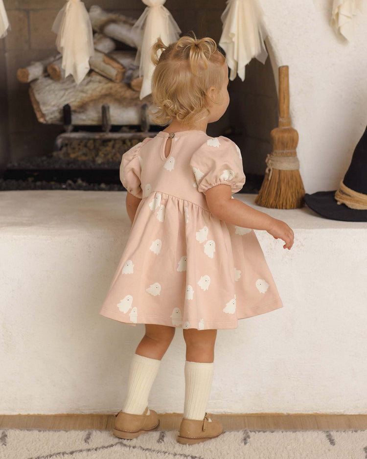 A young child with blonde hair in pigtails, dressed in the quincy mae darla dress in sweet ghost—a soft pink fleece style adorned with a charming ghost print—paired with cream socks and brown shoes, stands facing a fireplace decorated with hanging fabric and a broom on the hearth.