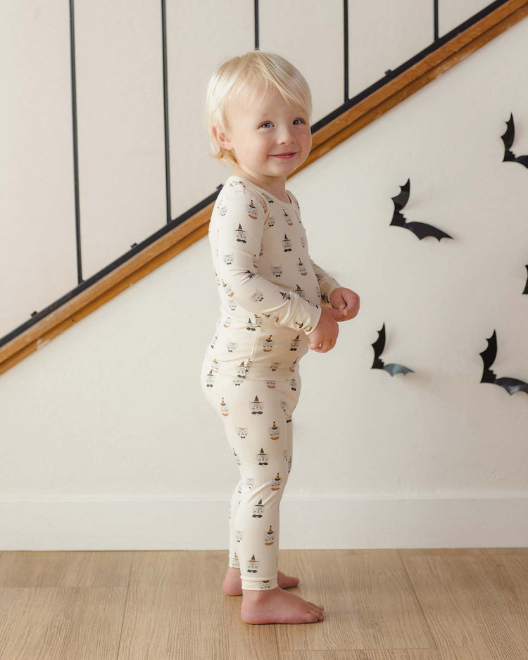 A young child with blonde hair wearing the Quincy Mae bamboo pajama set in costume kittens, a slim-fit long sleeve style, stands indoors near a staircase with black paper bats decorating the white wall behind them.