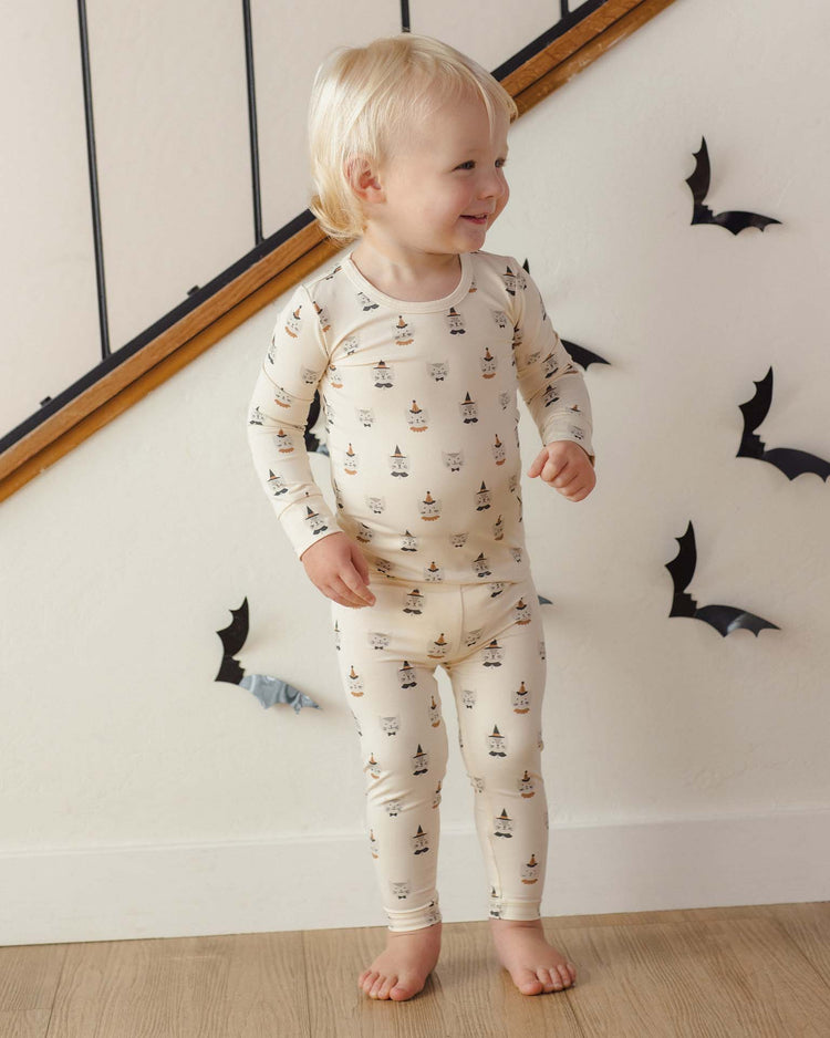A smiling toddler wearing the quincy mae bamboo pajama set in costume kittens stands indoors near stairs, surrounded by black paper bats on the wall.